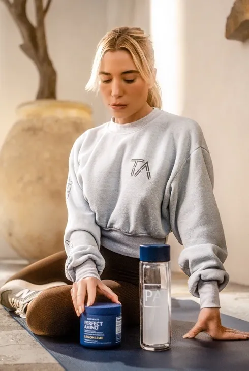 A woman in a sweatshirt sits on a yoga mat beside a jar of BodyHealth Perfect Amino and a water bottle